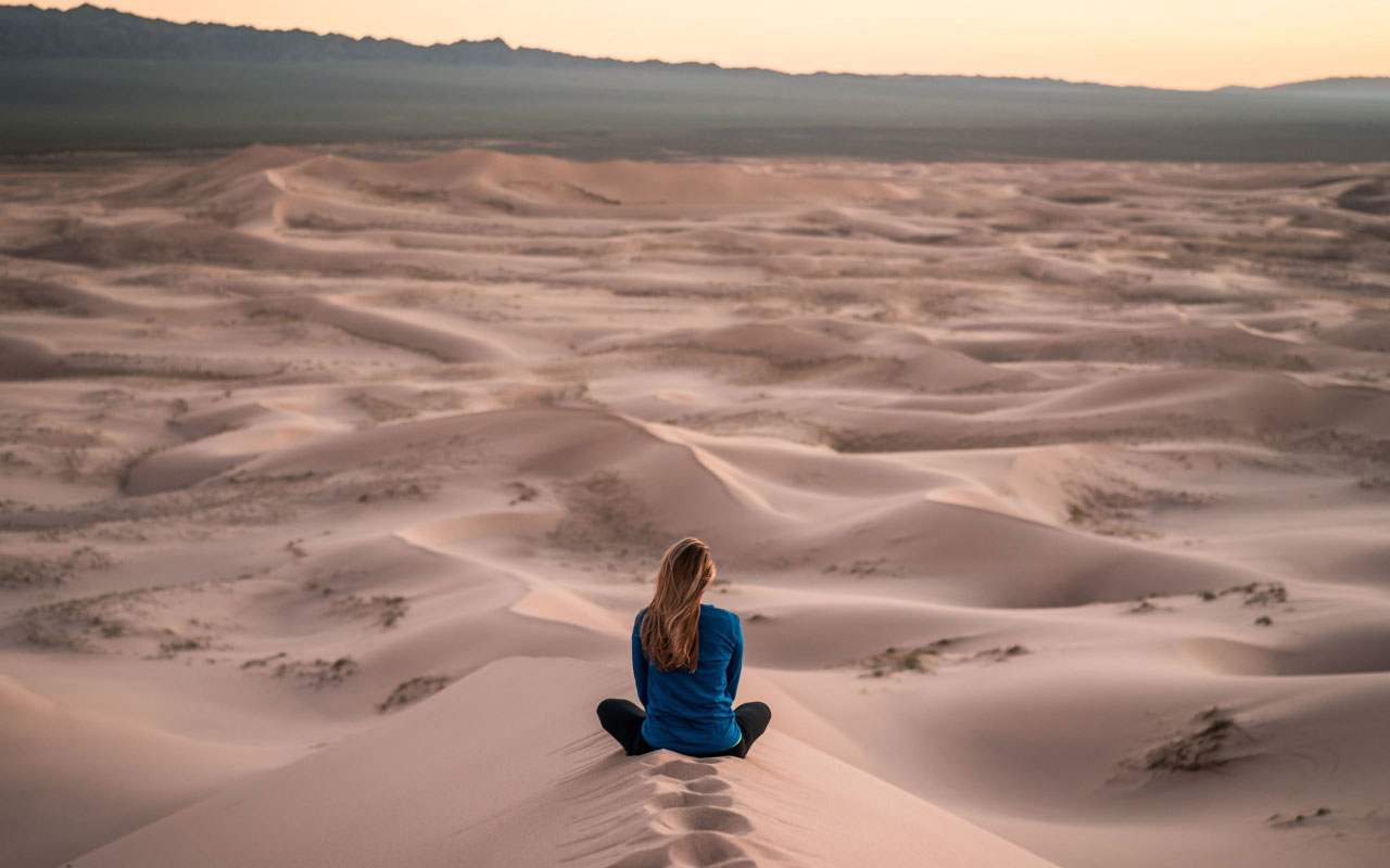 一个女人坐在沙漠中的沙丘上冥想。这可能是一个很好的引导可视化设置。