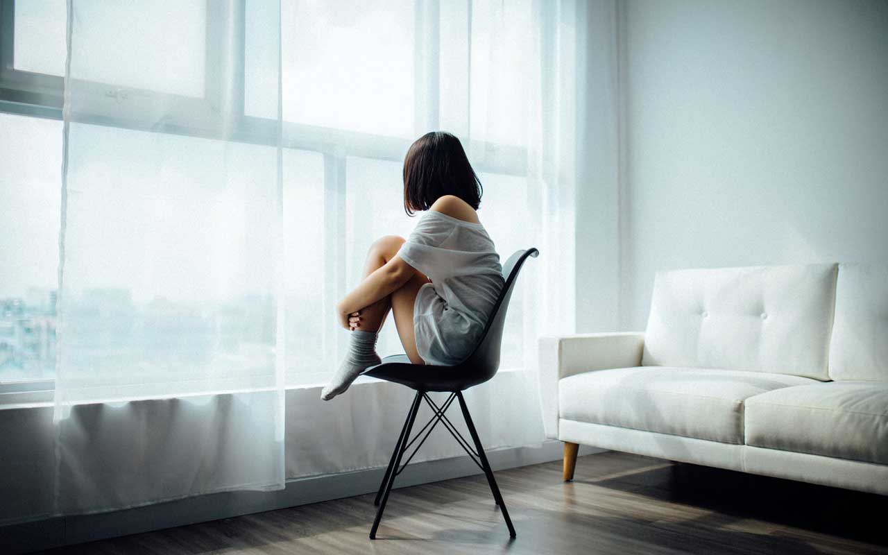一位妇女坐在椅子上，膝盖向上伸到胸前。她透过透明的窗帘和大玻璃窗向外张望。
