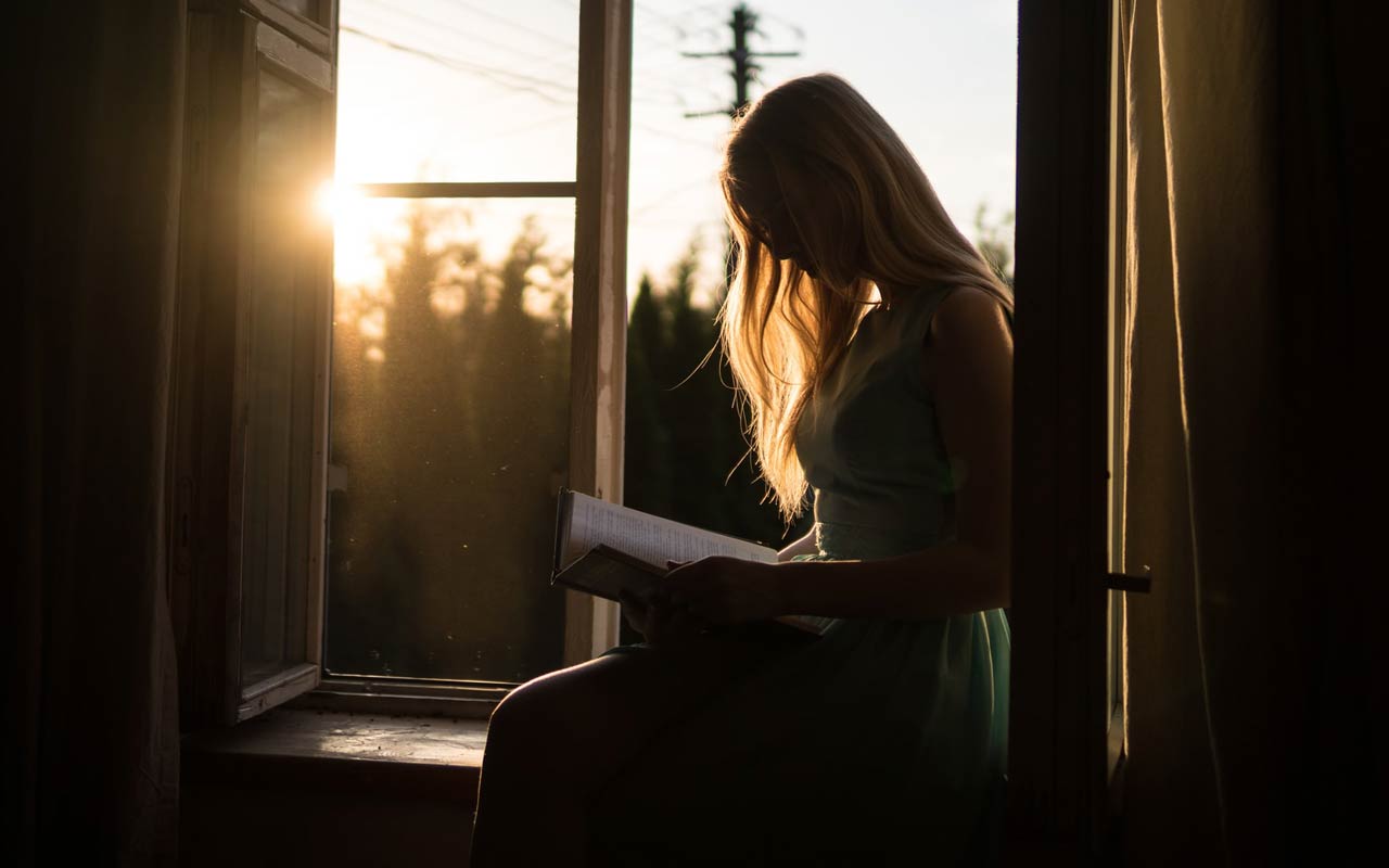 日落时分，一个女人坐在一扇开着的窗户旁，手里拿着一本书。
