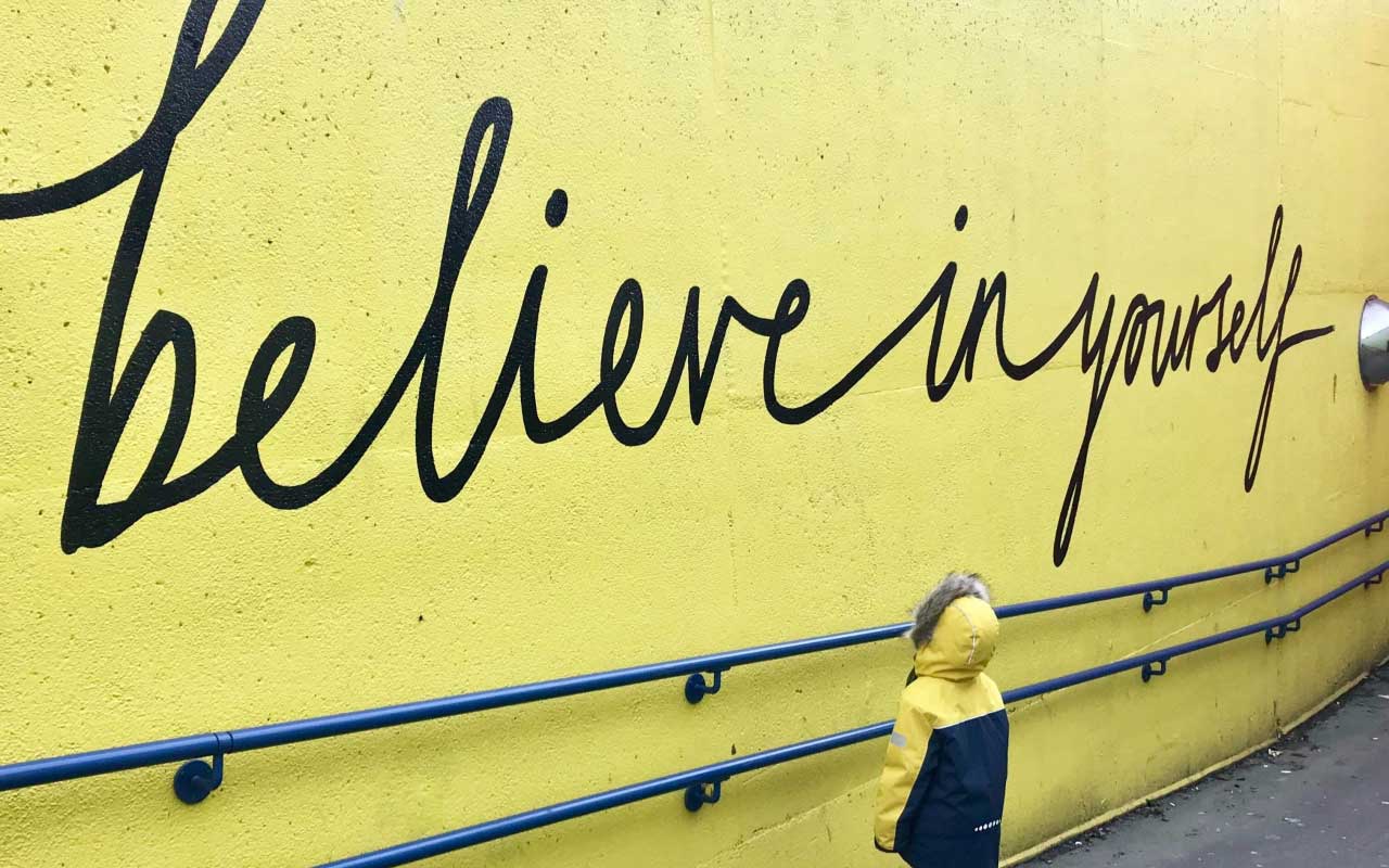 一个“相信自己”壁画绘在明亮的黄色墙上，有一个小孩看墙壁。