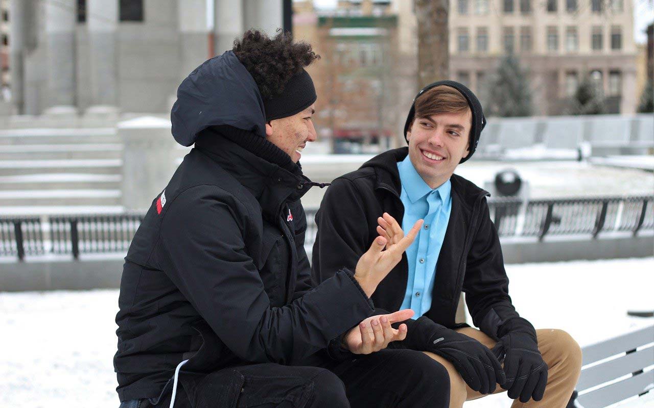 两个年轻人坐在替补席上，交谈。一个人在微笑着，而另一个会谈和使用手势。